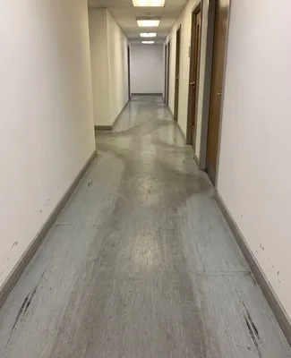 Before the machine wash, a long corridor of the office, a darker walkway in the middle of the light gray vinyl floor and shoe scratches, before the machine wash, protective tape is pasted on the door bases of the cabinets so that the washing water does not spill onto the carpets of the cabinets.