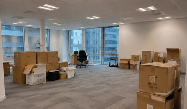 Stacks of moving boxes and a ladder in the office area under the large windows, during the cleaning after the move, the places of furniture marks left in the carpet are steamed up.