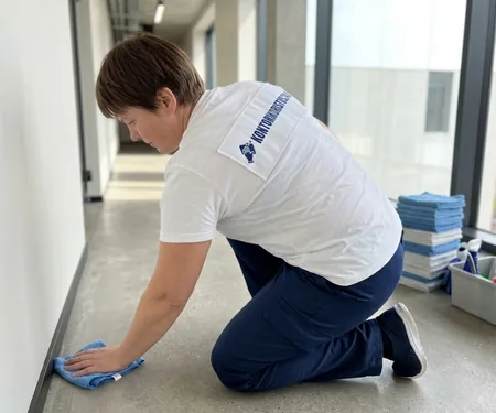 On his knees, a cleaner in a white shirt with a logo sweeps the floor next to the wall of the corridor with a cloth, the cleaner's initials are written on the checklist for each point made.