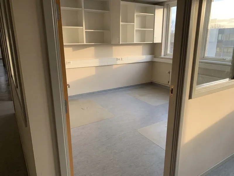 A small empty office with exposed white wall shelves and cable duct, lighter patches of gray vinyl floor showing the places of former desks and low sun shining through the window, items left behind by the tenant are boxed up and handed over to the owner.