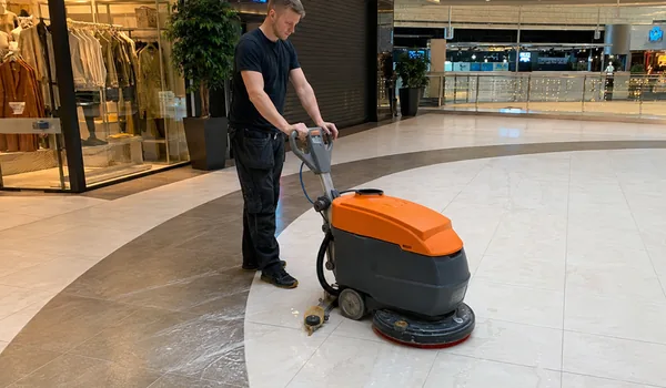 The operator controls the vacuum cleaner on the light stone floor of the shopping center between the shops, the wheels of the vacuum cleaner are made of non-marking rubber and do not leave marks on the stone.