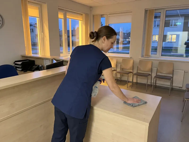 Koristaja tumesinises tööriietuses pühib lapiga kliiniku vastuvõtuleti, ooteruumi toolid on seina ääres reas ja akende taga on õhtu, kliiniku teravate jäätmete konteinerit koristaja ei ava ega tõsta.